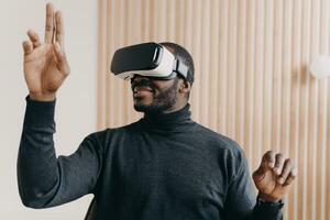 Smiling African American businessman in VR glasses, office setting, testing virtual reality goggles, impressed with 3D technology, interacting with virtual objects. photo