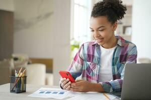 Mixed race girl uses mobile apps holding phone, distracted from learning at laptop. Gadget addiction photo