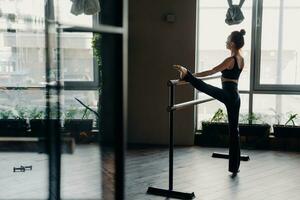 Slim red-haired ballerina stands in split position beside ballet barre in gym, light window background. Indoor sport and stretching showcased beautifully. photo