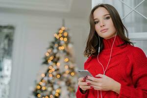 European girl with makeup, healthy skin, in oversized sweater, listens to audio book by Christmas tree. photo