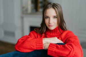 Brunette woman in loose red sweater, serious gaze, enjoys cozy day at home. Rest concept. photo