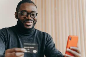 Positive African entrepreneur in glasses, paying online with credit card, ordering on cellphone, smiling millennial guy, using mobile banking app for transaction. photo