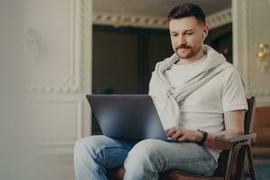 Happy young freelancer, casual clothes, earphones on, using laptop, working from modern home office, listening to music, focused on computer screen. photo