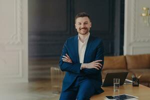 Satisfied bearded businessman in formal attire, stands confidently in office, arms folded, smiling pleasantly, content with work results. photo