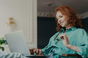 Redhead woman focused, happily working on remote job from modern apartment, preparing web publication. Eyeglasses in hand. photo