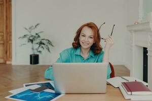Happy redhead woman in coworking space works on laptop, holds spectacles, browses web, communicates online. Blue shirt, paper documents. photo