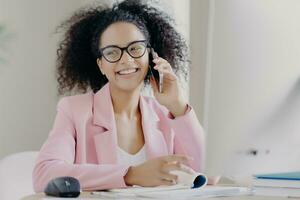 Happy curly-haired woman in rosy suit, glasses, enjoys phone talk with colleague, tech-filled break at desktop. photo