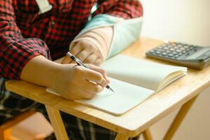 Hands of a male student were writing Mathematics homework in notebook with the calculator on table and His right arm was put in a splint. photo