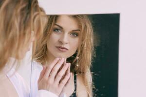 hermosa caucásico rubia joven mujer en blanco camisa en pie por el espejo. salud cuidado, belleza, mental cuestiones foto