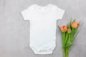 White baby girl or boy bodysuit mockup flat lay with orange tulips flowers decoration on the gray concrete background. Design onesie template, print presentation mock up. Top view. photo