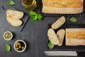 Fresh italian ciabatta bread with herbs, olive oil, black and green olives, basil leaves and pesto sauce on dark background. Top view. Copy space. photo