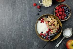 Bowl of homemade granola with yogurt and fresh berries on dark stone concrete background. Cranberry and apple crumble, crisp with almond flakes and oat. Top view. Copy space. photo