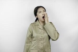 Portrait of sleepy Asian woman wearing a brown uniform and covering her mouth with a hand while yawning. Isolated image on white background photo