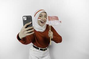 un sonriente asiático musulmán mujer vistiendo rojo parte superior y blanco hiyab, participación su teléfono y bandera, aislado por blanco antecedentes. de indonesia independencia día concepto foto