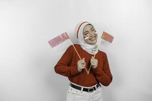 un joven asiático musulmán mujer con un contento exitoso expresión vistiendo rojo parte superior y blanco hijab mientras participación de indonesia bandera, aislado por blanco antecedentes. de indonesia independencia día concepto. foto