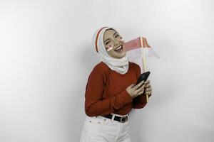 un sonriente asiático musulmán mujer vistiendo rojo parte superior y blanco hiyab, participación su teléfono y bandera, aislado por blanco antecedentes. de indonesia independencia día concepto foto