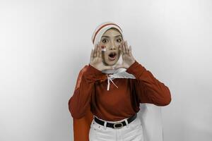 joven hermosa musulmán mujer vistiendo un rojo parte superior y blanco hijab es participación de indonesia bandera y gritos y gritando ruidoso con un mano en su boca. de indonesia independencia día concepto. foto