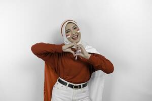 Happy smiling Indonesian muslim woman holding Indonesia's flag and showing love sign to celebrate Indonesia Independence Day isolated over white background. photo