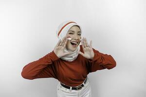 Young beautiful muslim woman wearing a red top and white hijab is shouting and screaming loud with a hand on her mouth. Indonesia's independence day concept. photo