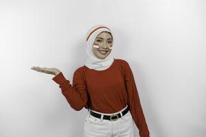 A happy Asian muslim woman wearing red top and white hijab, and pointing copy space beside her, isolated by white background. Indonesia's independence day photo