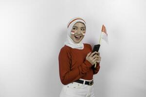 un sonriente asiático musulmán mujer vistiendo rojo parte superior y blanco hiyab, participación su teléfono y bandera, aislado por blanco antecedentes. de indonesia independencia día concepto foto