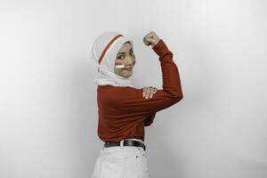 emocionado asiático musulmán mujer vistiendo un rojo parte superior y blanco hijab demostración fuerte gesto por levantamiento su brazos y músculos sonriente con orgullo de indonesia independencia día concepto. foto