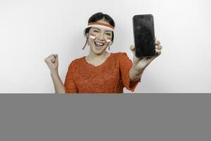 A young Asian woman with a happy successful expression while holding her phone and wearing red kebaya, flag headband isolated by white background. Indonesia's independence day concept. photo