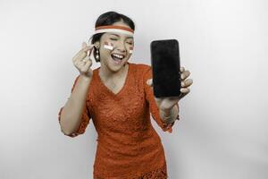 A happy Asian woman wearing red kebaya and headband, showing her phone, and gesturing love sign by her fingers showing tender feeling, isolated by white background. Indonesia's independence day photo