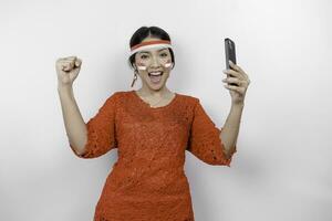 A young Asian woman with a happy successful expression while holding her phone and wearing red kebaya, flag headband isolated by white background. Indonesia's independence day concept. photo