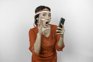 joven hermosa mujer vistiendo un rojo kebaya es participación su teléfono mientras gritos y gritando ruidoso con un mano en su boca. de indonesia independencia día concepto. foto
