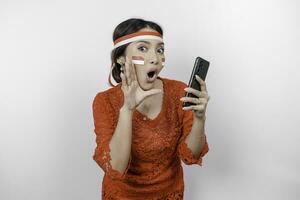 Young beautiful woman wearing a red kebaya is holding her phone while shouting and screaming loud with a hand on her mouth. Indonesia's independence day concept. photo