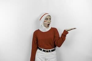 A shocked Asian muslim woman wearing white hijab and red top, and pointing copy space beside her, isolated by white background. Indonesia's independence day concept photo