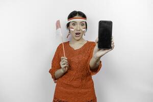 A portrait of a shocked Asian woman wearing red kebaya and headband, showing her phone while her mouth wide open, isolated by white background. Indonesia's independence day concept photo