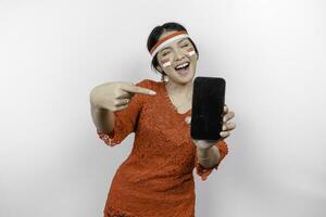 A portrait of a smiling Asian woman wearing red kebaya and headband and showing her phone, isolated by white background. Indonesia's independence day concept photo