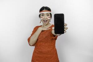 A portrait of a shocked Asian woman wearing red kebaya and headband, showing her phone while her mouth wide open, isolated by white background. Indonesia's independence day concept photo