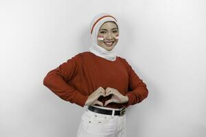 un joven asiático mujer vistiendo un rojo parte superior y blanco hijab siente nacionalismo, formas corazón gesto expresa su amor para Indonesia. de indonesia independencia día concepto. foto