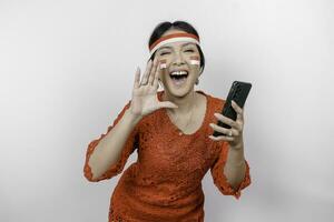 Young beautiful woman wearing a red kebaya is holding her phone while shouting and screaming loud with a hand on her mouth. Indonesia's independence day concept. photo