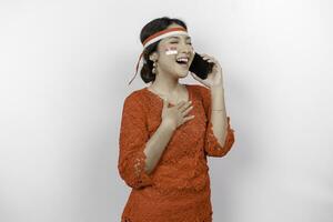 Happy mindful thankful young Asian woman wearing red kebaya and holding phone and hand on chest smiling isolated on white background feeling no stress, photo