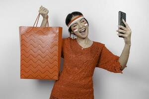 An excited Asian woman wearing red kebaya holding a shopping bag and her smartphone, studio shot isolated on white background. Indonesia's Independence day concept photo