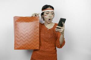 A shocked Asian woman wearing red kebaya holding a shopping bag and her smartphone, studio shot isolated on white background. Indonesia's Independence day concept photo