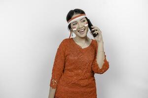 A portrait of a smiling Asian woman wearing red kebaya and headband and holding her phone, isolated by white background. Indonesia's independence day concept photo