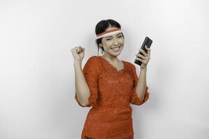 A young Asian woman with a happy successful expression while holding her phone and wearing red kebaya, flag headband isolated by white background. Indonesia's independence day concept. photo