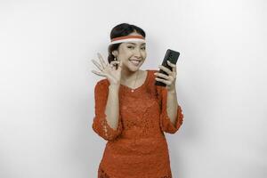 A cheerful Asian woman wearing red kebaya and headband, holding her phone while gesturing OK sign with her fingers, isolated by white background. Indonesia's independence day concept photo