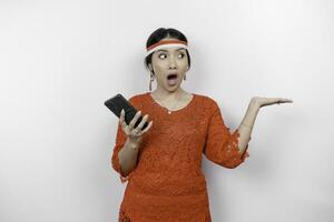 A portrait of a shocked Asian woman wearing red kebaya and headband, holding her phone, and pointing copy space beside her, isolated by white background. Indonesia's independence day concept photo