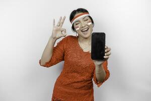 un alegre asiático mujer vistiendo rojo kebaya y venda, demostración su teléfono mientras gesticulando Okay firmar con su dedos, aislado por blanco antecedentes. de indonesia independencia día concepto foto
