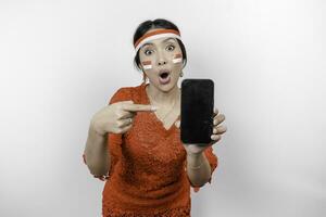 A portrait of a shocked Asian woman wearing red kebaya and headband, showing her phone while her mouth wide open, isolated by white background. Indonesia's independence day concept photo