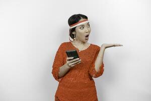 A portrait of a shocked Asian woman wearing red kebaya and headband, holding her phone, and pointing copy space beside her, isolated by white background. Indonesia's independence day concept photo