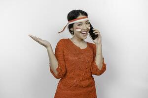 A happy Asian woman wearing red kebaya and headband, holding her phone, and pointing copy space beside her, isolated by white background. Indonesia's independence day photo