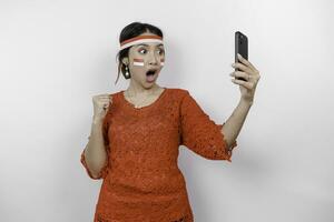 A young Asian woman with a happy successful expression while holding her phone and wearing red kebaya, flag headband isolated by white background. Indonesia's independence day concept. photo