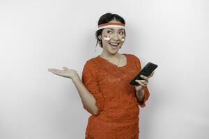 A happy Asian woman wearing red kebaya and headband, holding her phone, and pointing copy space beside her, isolated by white background. Indonesia's independence day photo